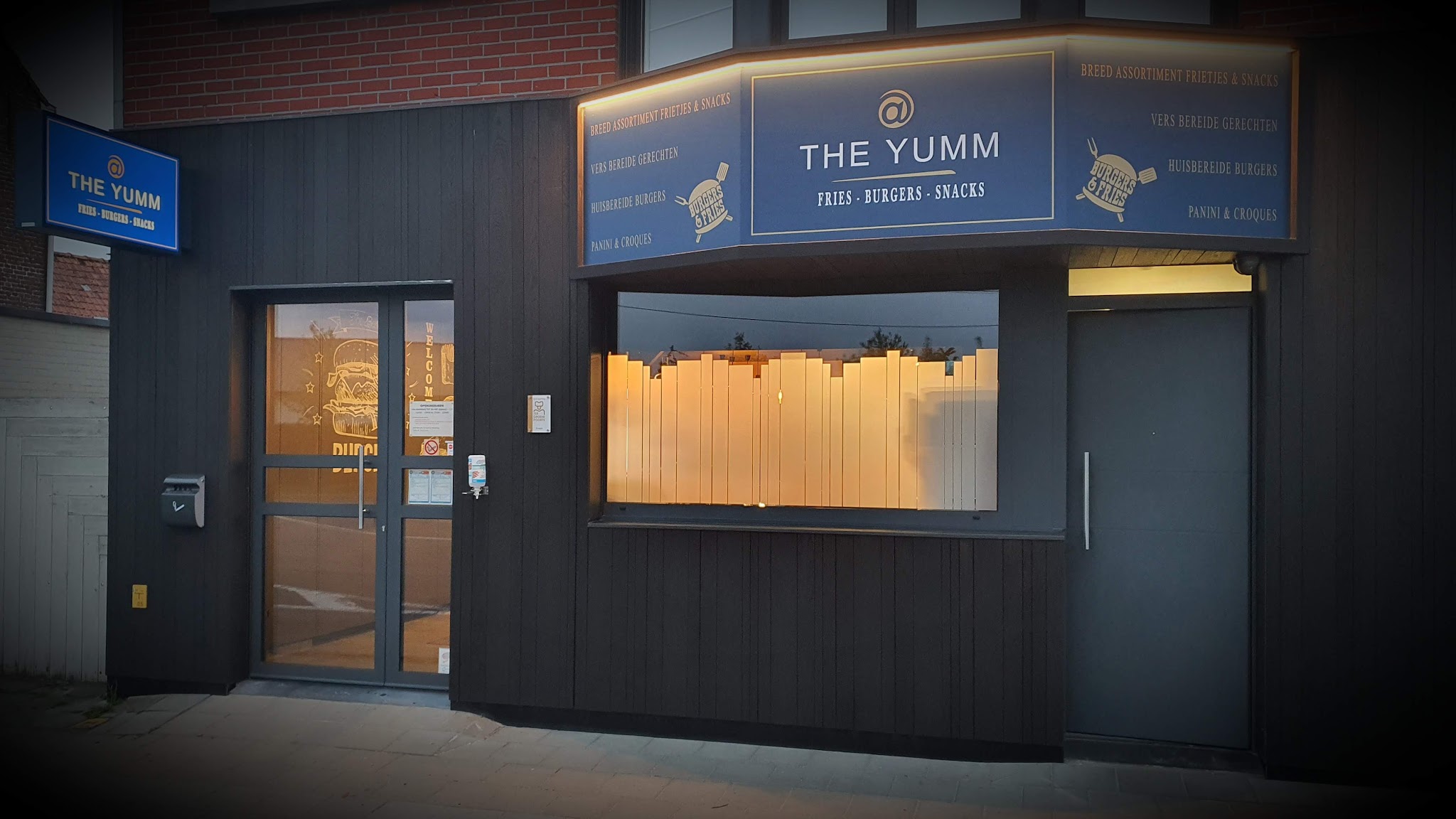 De gevel van @ The Yumm in Koolskamp bij valavond, met het verlichte blauwe uithangbord